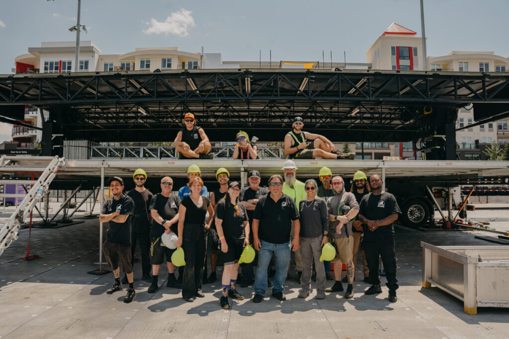 About twenty ShadowMonkey crew in black crew shirts in front of a trailer-mounted outdoor stage, many holding hi-vis hard hats, with three more seated on the deck above.