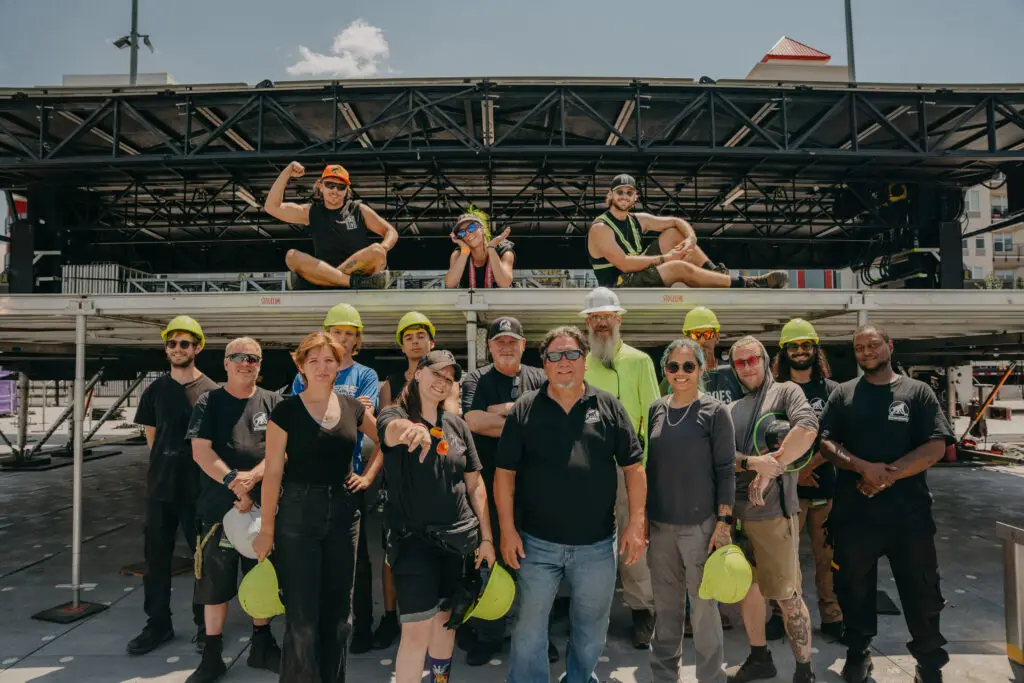 A ShadowMonkey stage crew of about eighteen gathered in front of a rigged outdoor stage roof, most in hi-vis hard hats and crew shirts.